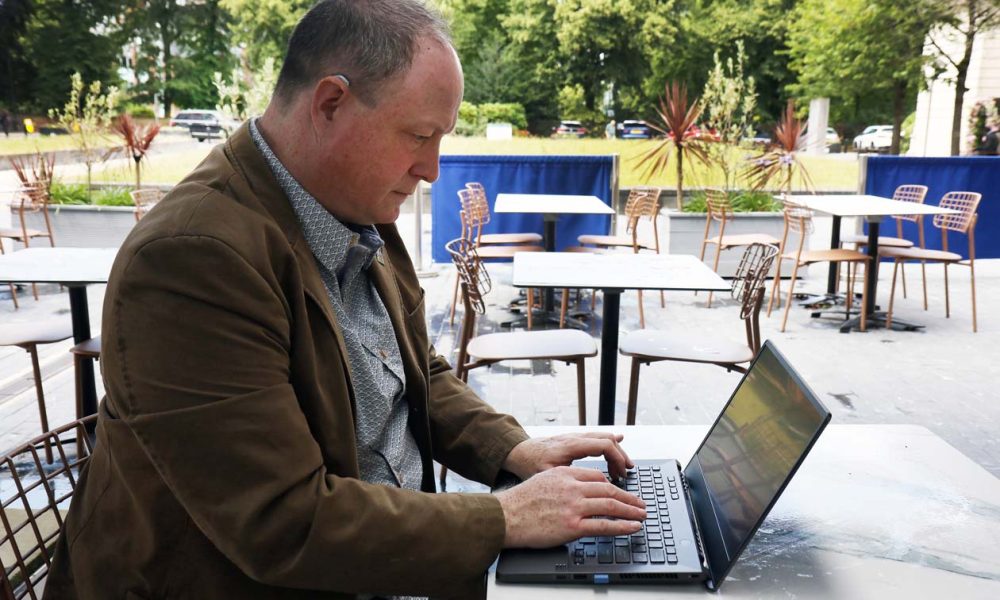Would you like us to review your Auracast-enabled product? Phil Creighton at an open air cafe typing a review on a laptop