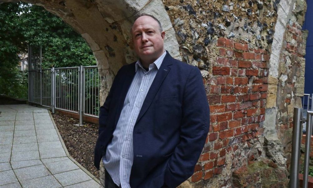Phil Creighton leaning against an archway that was once part of Reading Abbey. There is red brickwork behind the arch. On the left there is a footpathy and trees and metal fencing can be seen behind it.
