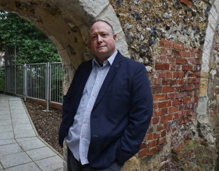 Phil Creighton leaning against an archway that was once part of Reading Abbey. There is red brickwork behind the arch. On the left there is a footpathy and trees and metal fencing can be seen behind it.