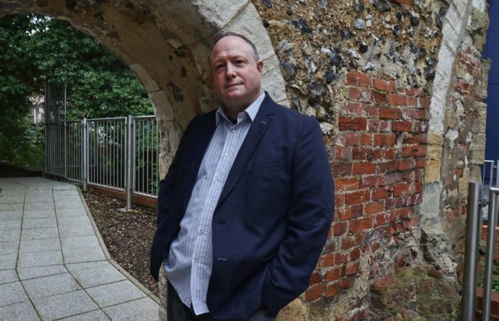 Phil Creighton leaning against an archway that was once part of Reading Abbey. There is red brickwork behind the arch. On the left there is a footpathy and trees and metal fencing can be seen behind it.