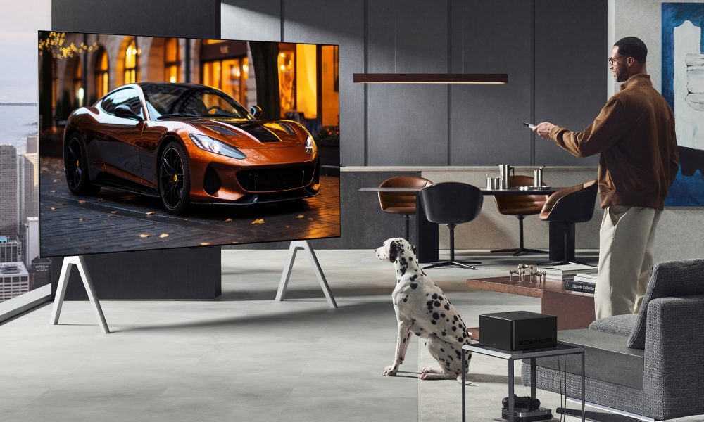 A man watching an LG television in a lounge with a Dalmatian next to him. The man is standing up and has a remote control in his hands. In the backrgound there is a kitchen. The television is massive and has a flash car on the screen.