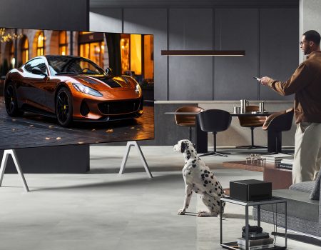 A man watching an LG television in a lounge with a Dalmatian next to him. The man is standing up and has a remote control in his hands. In the backrgound there is a kitchen. The television is massive and has a flash car on the screen.