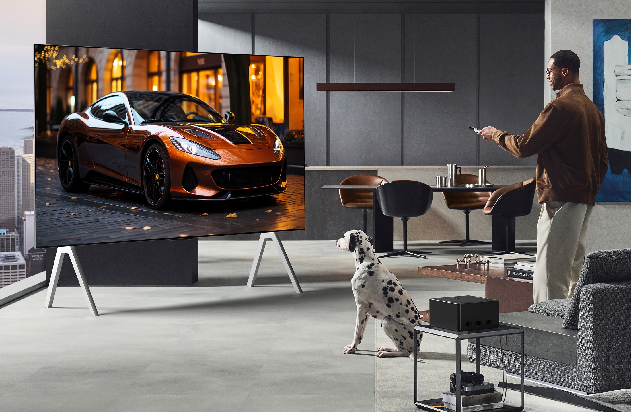 A man watching an LG television in a lounge with a Dalmatian next to him. The man is standing up and has a remote control in his hands. In the backrgound there is a kitchen. The television is massive and has a flash car on the screen.
