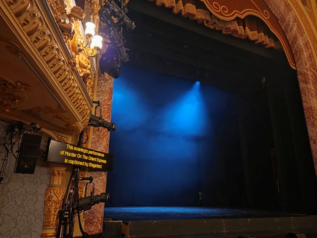 The captioning machine can be seen at the side of the Everyman Cheltenham stage. Two blue spotlights are shining on the empty stage, while on the far left the upper tier box is visible