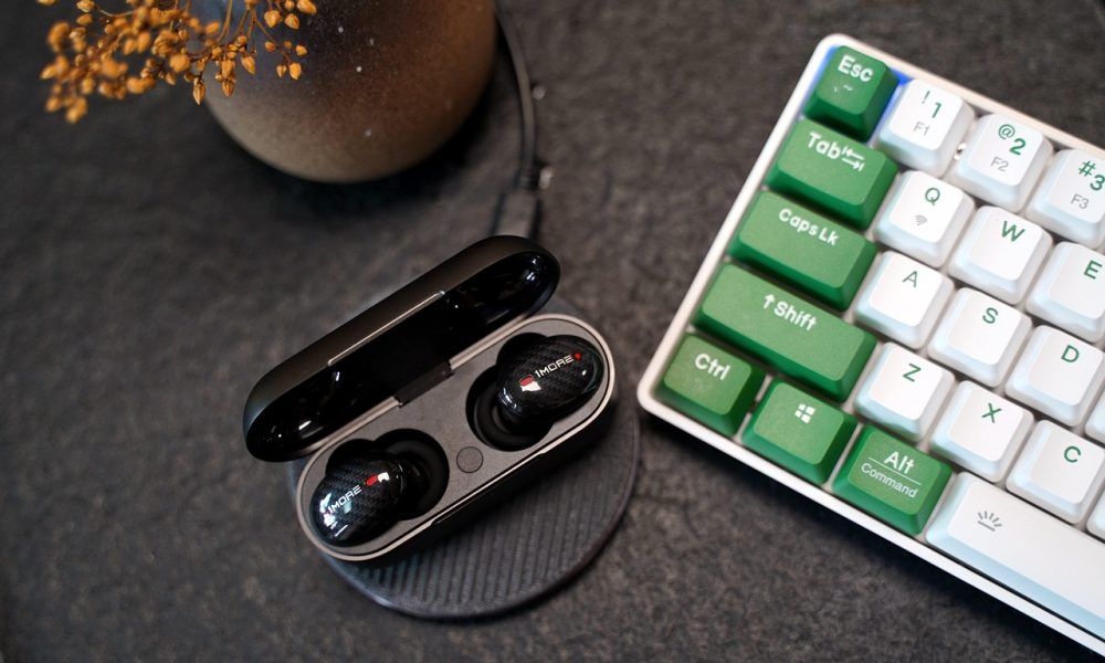 A pair of Bluetooth earbuds in their case next to a keyboard. Some of the command keys are in green. In the top left hand corner is a plant pot.