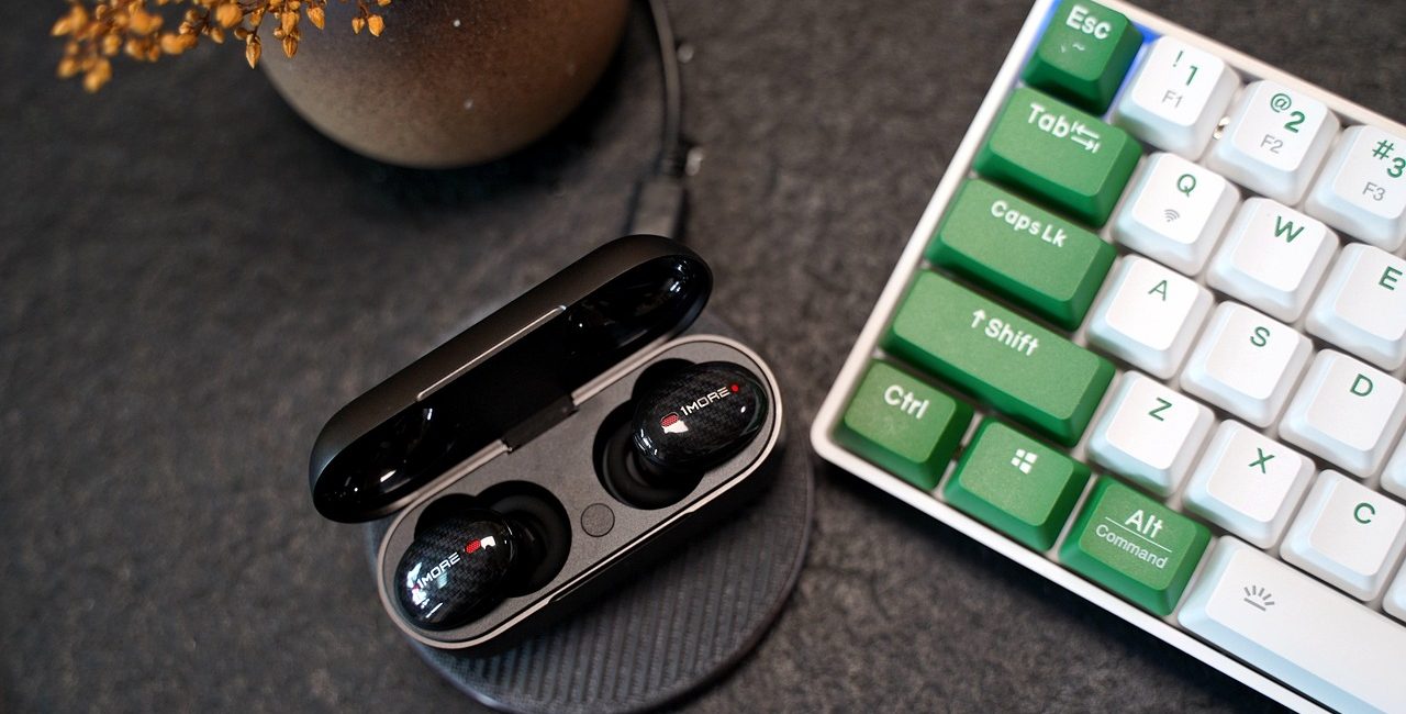 A pair of Bluetooth earbuds in their case next to a keyboard. Some of the command keys are in green. In the top left hand corner is a plant pot.
