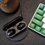 A pair of Bluetooth earbuds in their case next to a keyboard. Some of the command keys are in green. In the top left hand corner is a plant pot.
