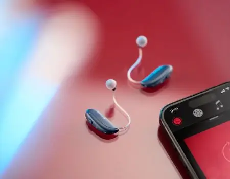 The ReSound Vivia hearing aids on a red background. On the right hand side of the image is a mobile phone showing the ReSound app