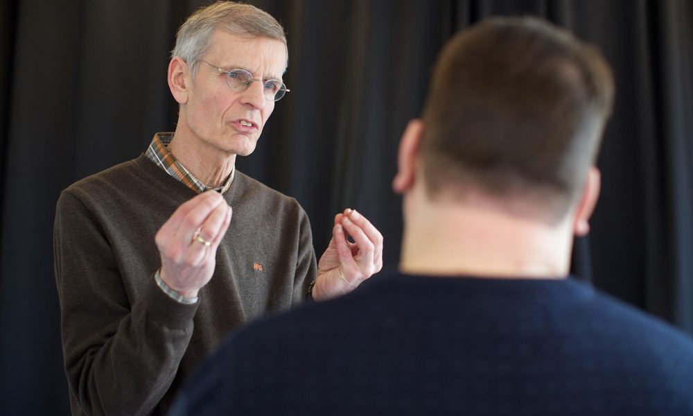 A stock image of a man making a point to another person with their back to us. The person speaking is raising their pinched hands to illustrate something. Bluetooth SIG is holding an Auracast Advocates seminar this