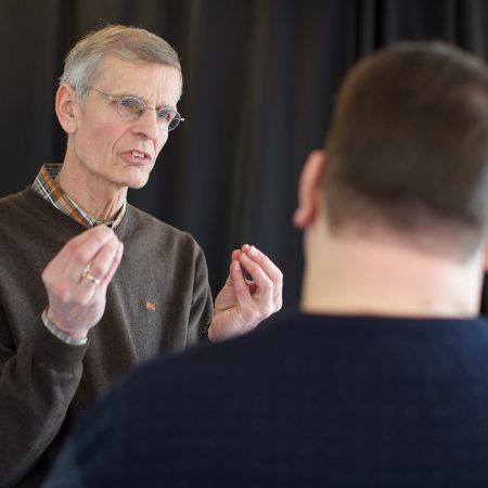 A stock image of a man making a point to another person with their back to us. The person speaking is raising their pinched hands to illustrate something. Bluetooth SIG is holding an Auracast Advocates seminar this