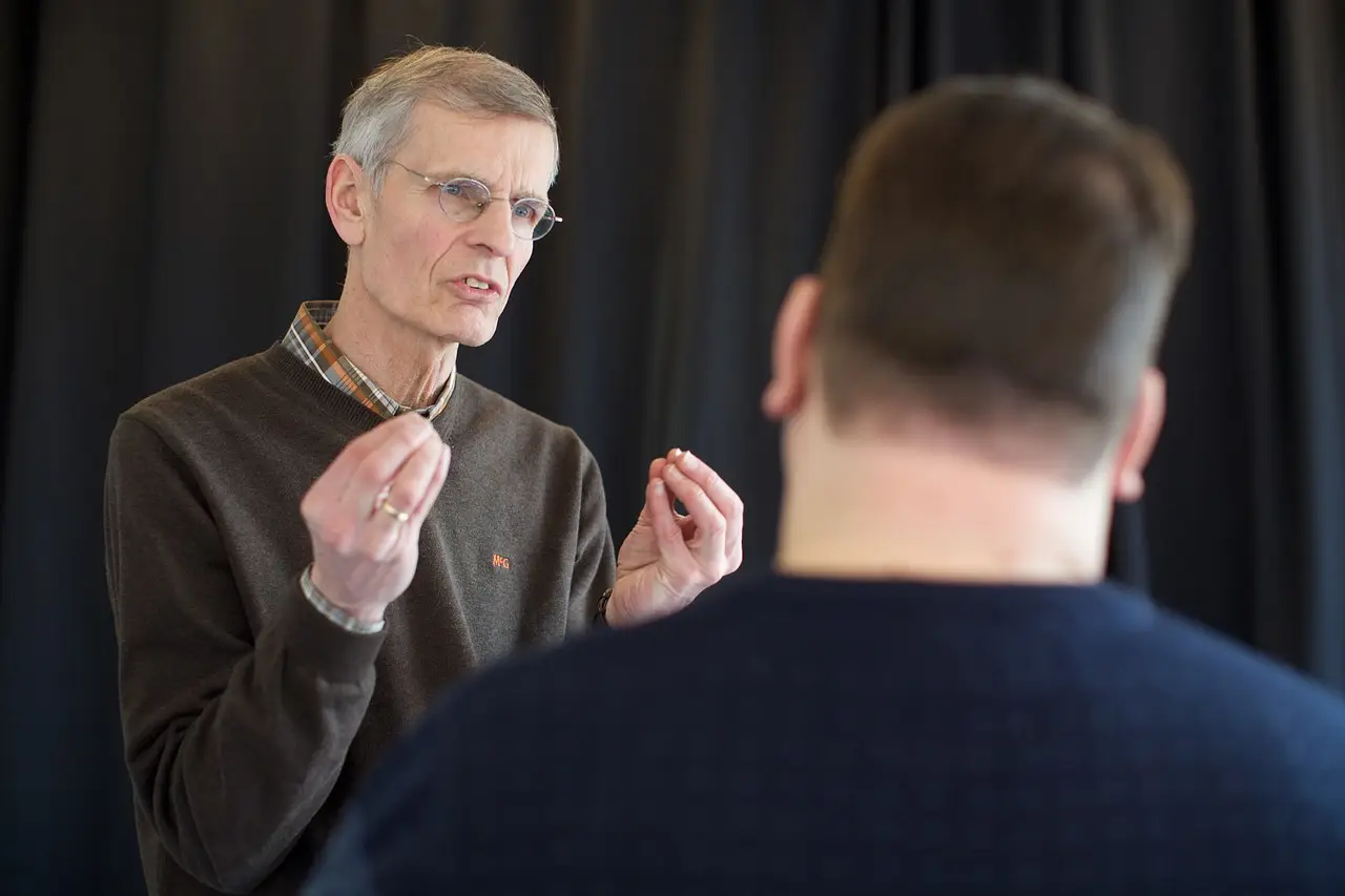 A stock image of a man making a point to another person with their back to us. The person speaking is raising their pinched hands to illustrate something. Bluetooth SIG is holding an Auracast Advocates seminar this