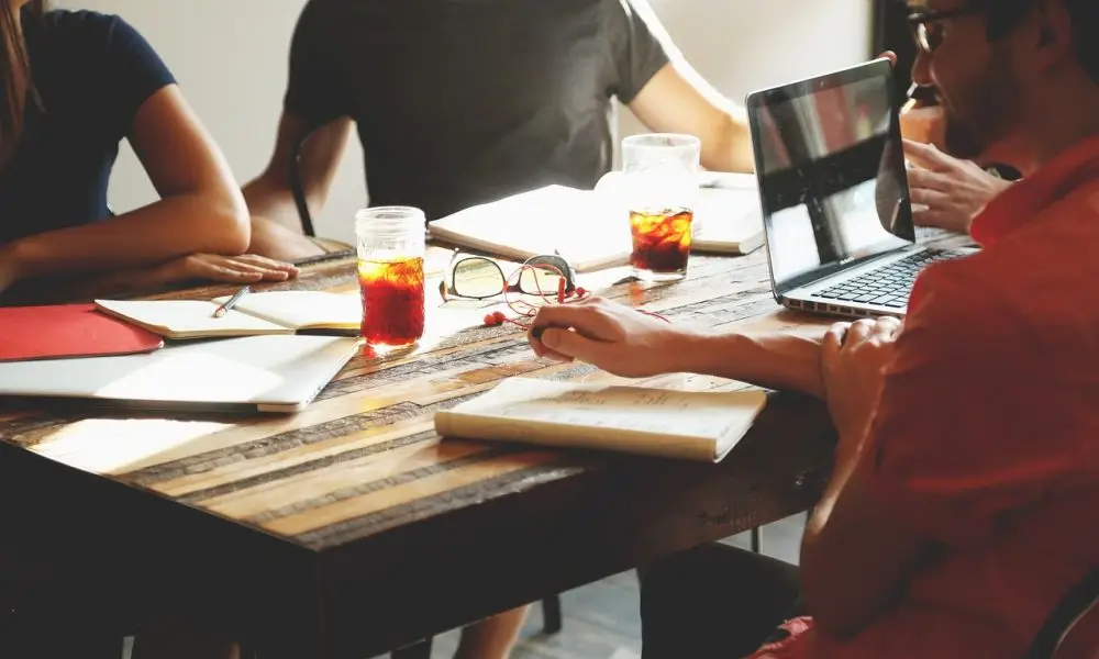 People sitting around a table at a business meeting. We cannot see faces, only torsos and arms. There are drinks, paperework and a laptop on the table. The picture helps illustrate a meeting where Auracast Advocates are sharing information about the Bluetooth technology