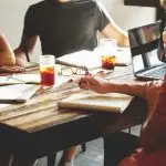 People sitting around a table at a business meeting. We cannot see faces, only torsos and arms. There are drinks, paperework and a laptop on the table. The picture helps illustrate a meeting where Auracast Advocates are sharing information about the Bluetooth technology
