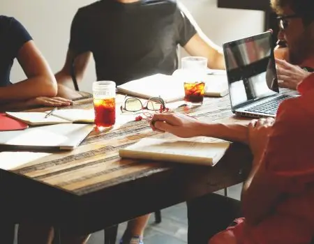 People sitting around a table at a business meeting. We cannot see faces, only torsos and arms. There are drinks, paperework and a laptop on the table. The picture helps illustrate a meeting where Auracast Advocates are sharing information about the Bluetooth technology