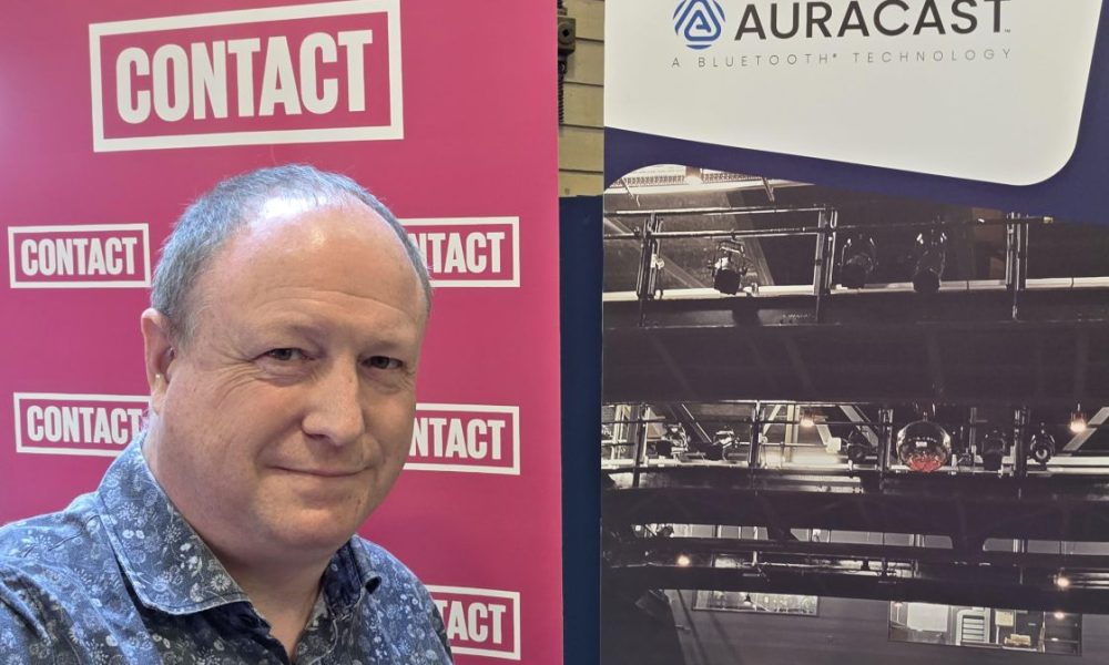 Phil Creighton standing next to a banner saying CONTACT and a banner with the Auracast Logo
