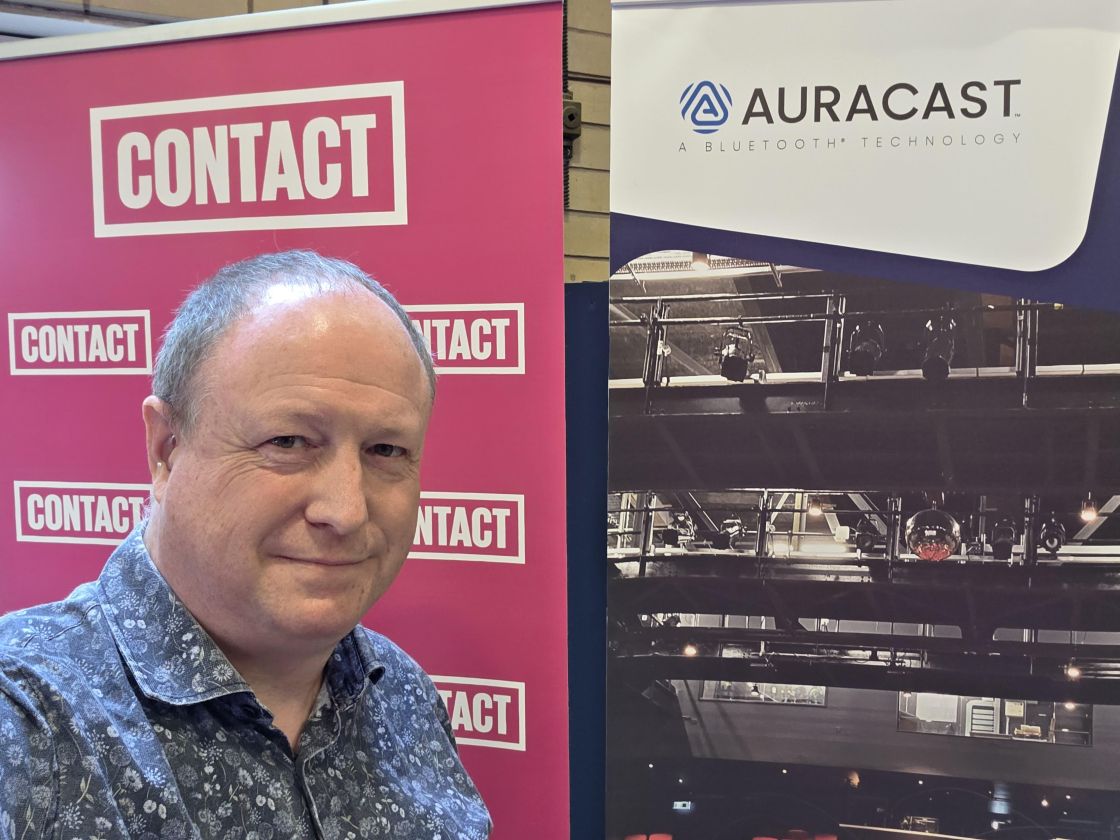 Phil Creighton standing next to a banner saying CONTACT and a banner with the Auracast Logo