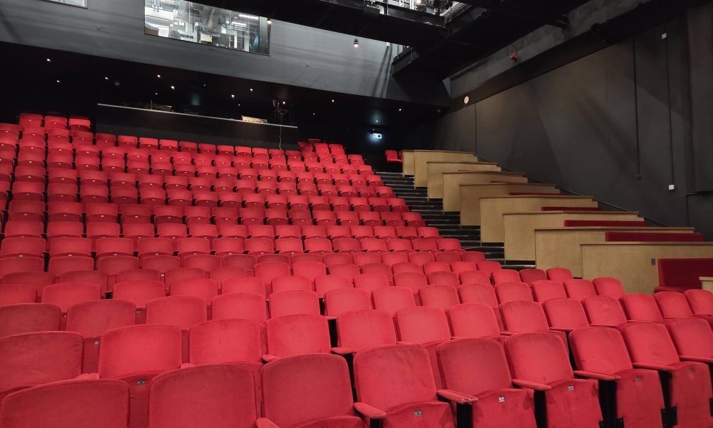 Auracast 2030 is a vision to see Auracast installed in theatres such as this one. Pictured are rows and rows of red seats in a theatre auditorium. The venue is empty