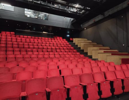 Auracast 2030 is a vision to see Auracast installed in theatres such as this one. Pictured are rows and rows of red seats in a theatre auditorium. The venue is empty