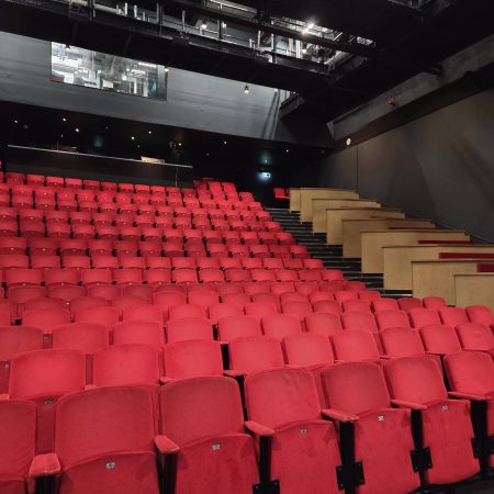 Auracast 2030 is a vision to see Auracast installed in theatres such as this one. Pictured are rows and rows of red seats in a theatre auditorium. The venue is empty