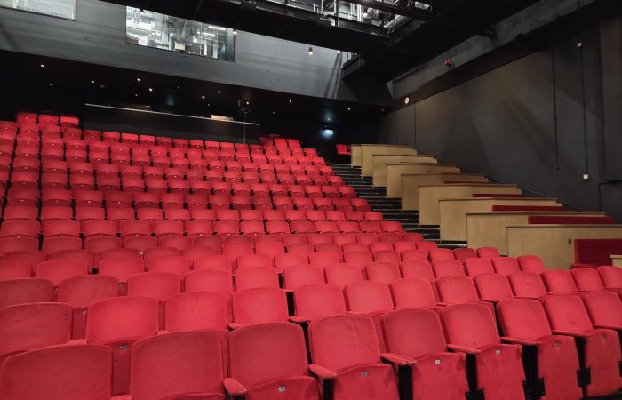 Auracast 2030 is a vision to see Auracast installed in theatres such as this one. Pictured are rows and rows of red seats in a theatre auditorium. The venue is empty
