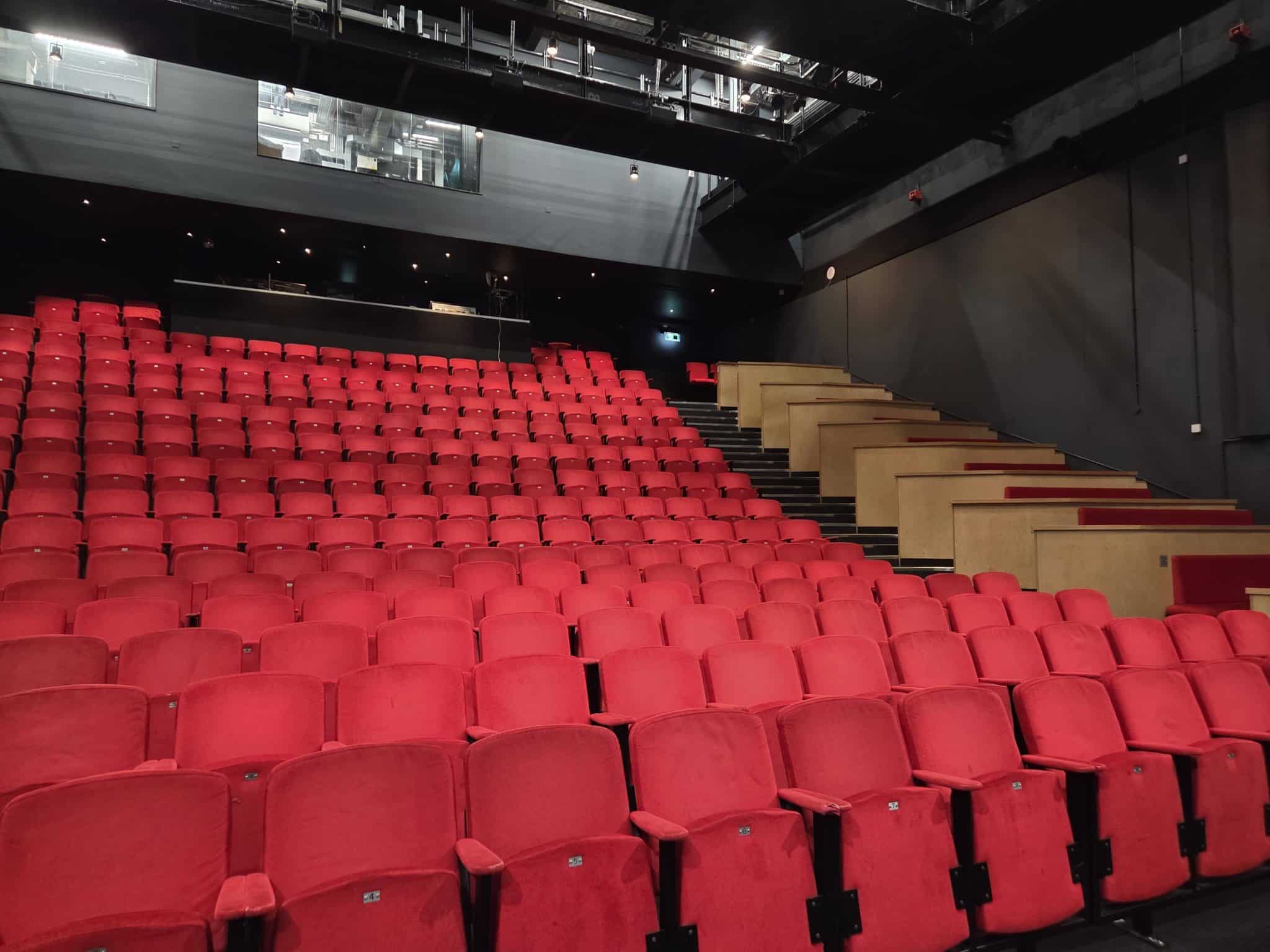 Auracast 2030 is a vision to see Auracast installed in theatres such as this one. Pictured are rows and rows of red seats in a theatre auditorium. The venue is empty
