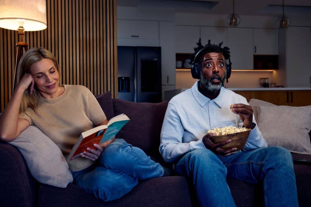 The Sennheiser RS 275 headphones and BTA1 transmitter can be paired for silent listening: The picture shows a woman sitting on a sofa reading a book, while a man is wearing headphones and enjoying popcorn while watching something on an unseen television