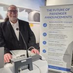 A man standing at a desk with an Auracast-enabled public address system in front of him. A banner is behind him