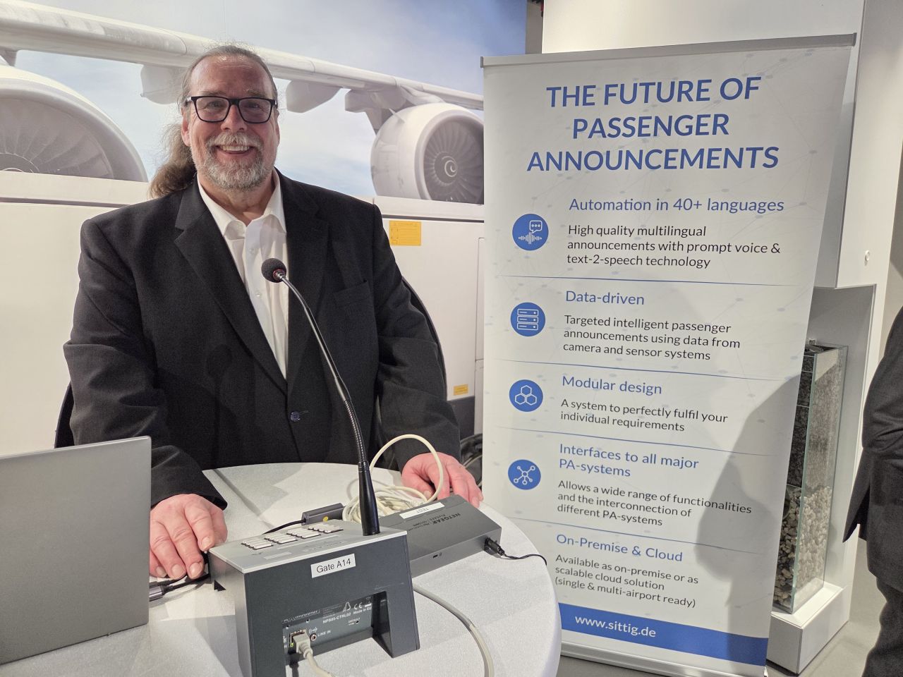 A man standing at a desk with an Auracast-enabled public address system in front of him. A banner is behind him